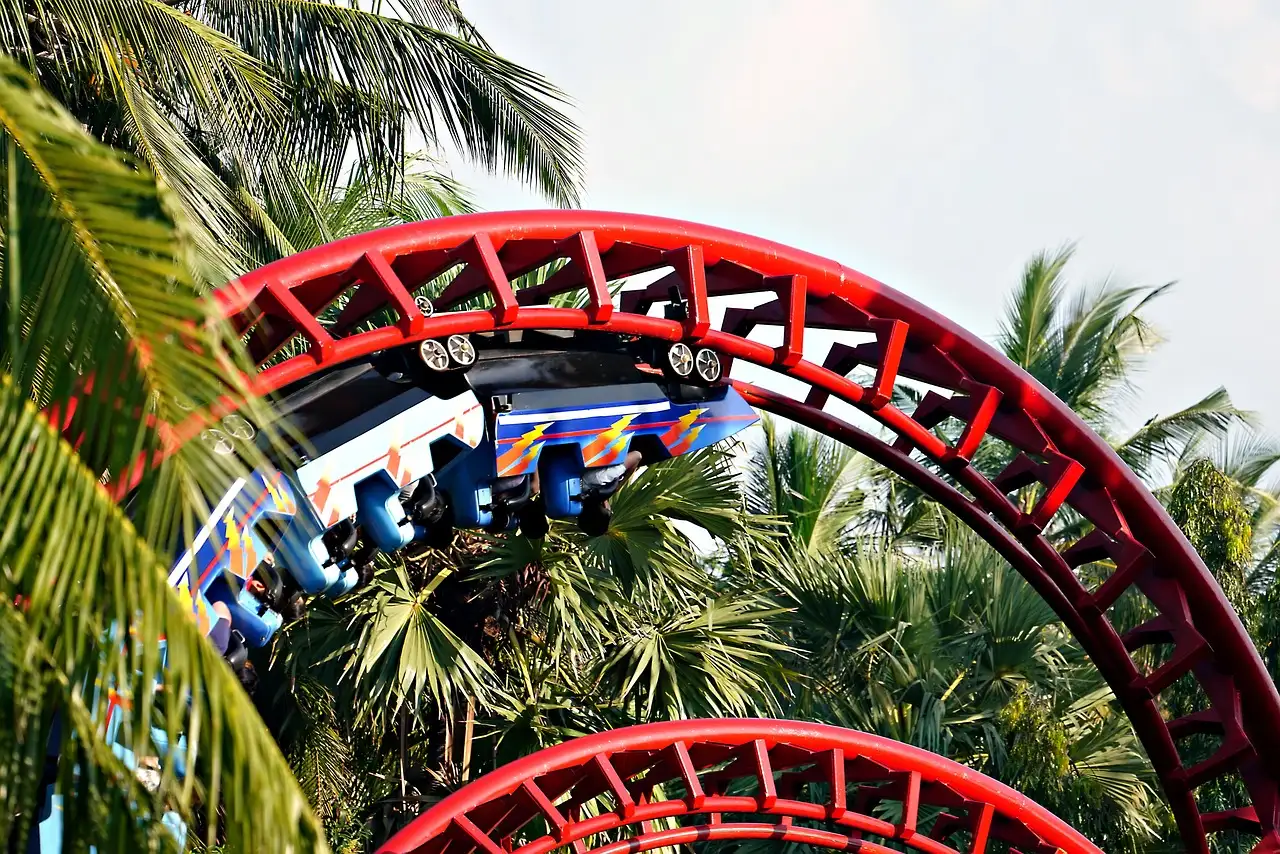 A red roller coaster track curving through palm trees, with a train of riders on it