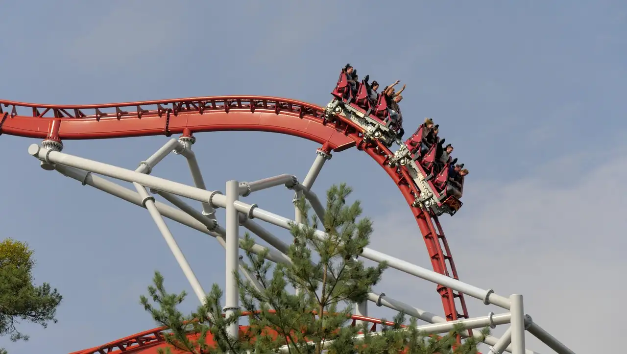 A red roller coaster train with riders coming over a curve above a pine tree