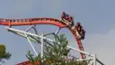 A red roller coaster train with riders coming over a curve above a pine tree