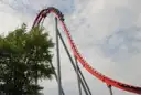 A red roller coaster train going over a tall hill against a cloudy sky