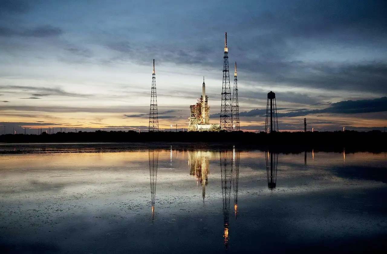 A rocket on its launch tower at sunrise, reflected in calm water