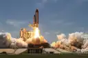 A space shuttle launching by day with white smoke billowing out