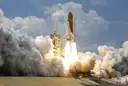 A space shuttle lifting off from a launch pad, with big clouds of smoke on both sides