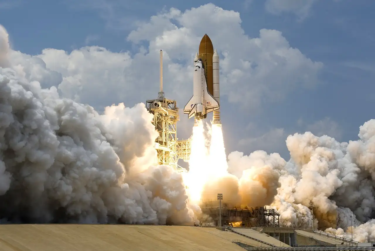 A space shuttle lifting off from a launch pad, with big clouds of smoke on both sides