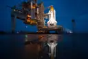 A space shuttle on a lit launch pad at dusk, reflected in wet ground