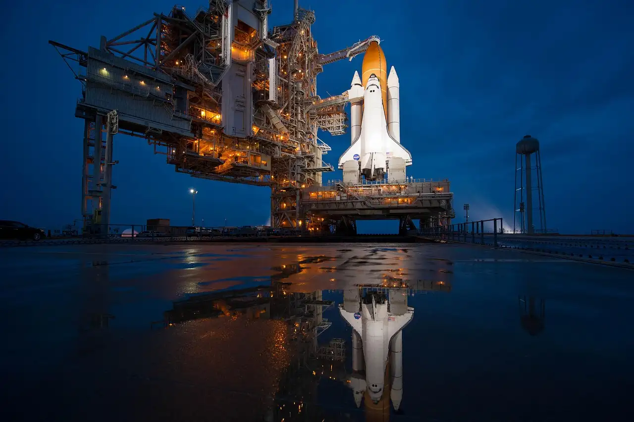 A space shuttle on a lit launch pad at dusk, reflected in wet ground