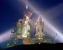 A space shuttle on its launch pad at night, lit by searchlight beams